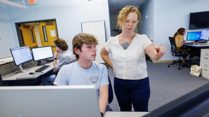 Katrina Jagodinsky stands next to student Luke McDermott, seated at a computer, with two student on computers in the background.