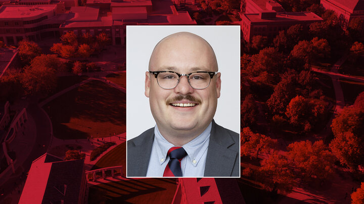 Color portrait of Julian Gillilan, doctoral candidate in the University of Nebraska–Lincoln's Department of History, on a red campus background. He is wearing a dark gray sport coat over a light blue dress shirt and red-and-blue striped tie, with glasses.